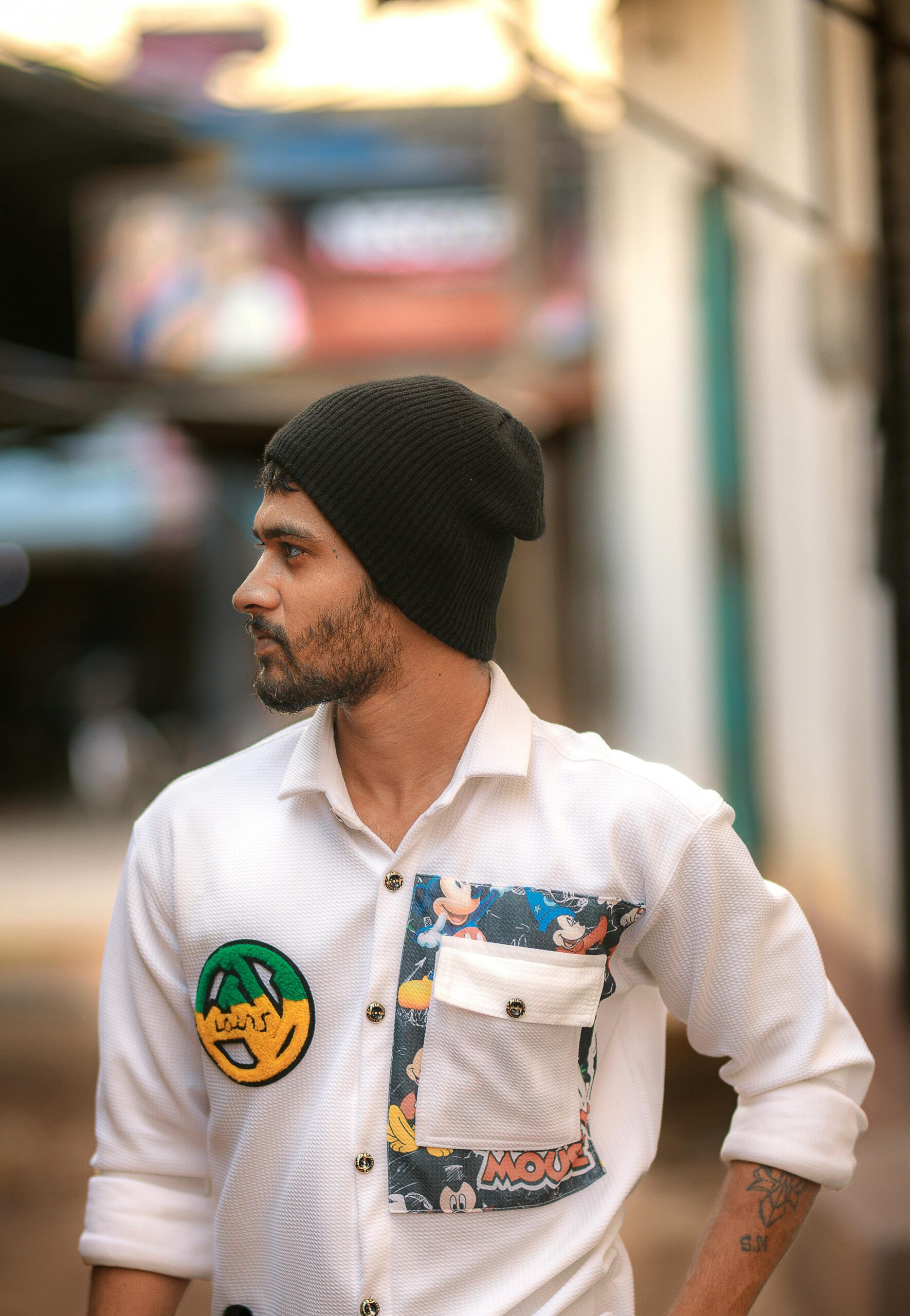 A stylish young man wearing a casual outfit with playful patches, standing in an urban area.
